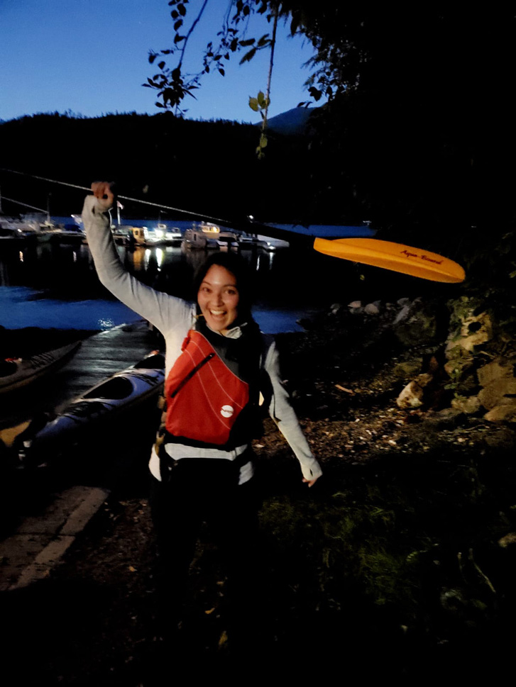 Editor Alyssa at night raising a paddle in the air for a kayak by the water