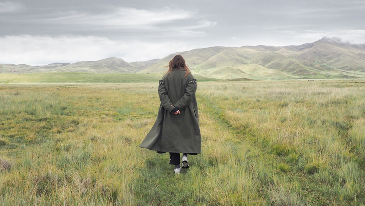 Woman back turned in trenchcoat staring at hill landscape