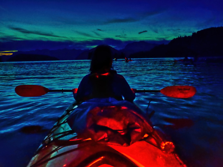 From behind a kayak, woman rowing at night in kayak on water
