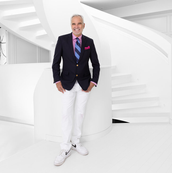 Full body standing Paul Lavoie in a dark suit jacket and white pants in front of white staircase and white room