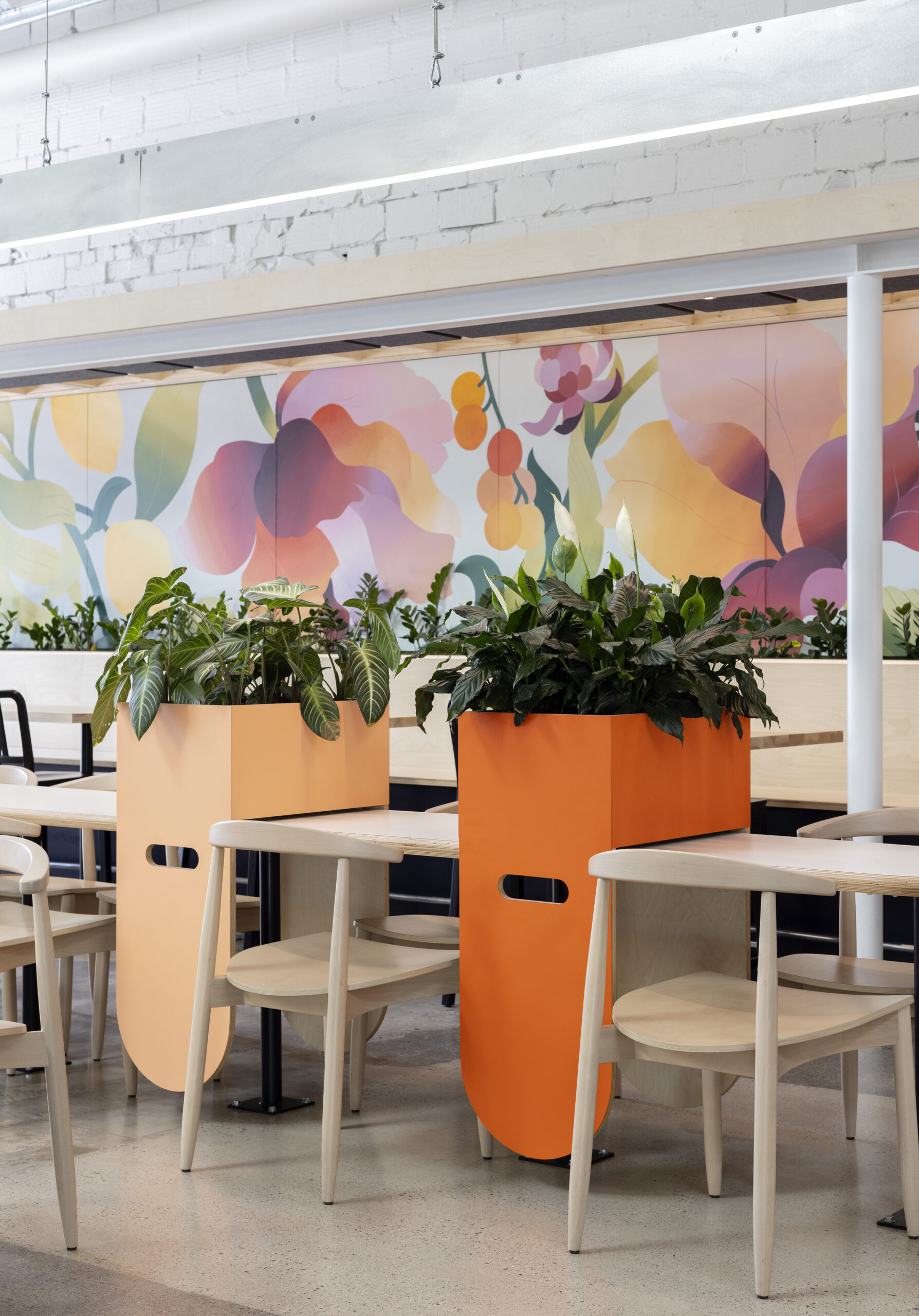 Close-up view of a vibrant and inviting café corner featuring sleek wooden tables, chairs, and striking orange planters filled with lush greenery, set against a backdrop of a large, colourful floral mural.