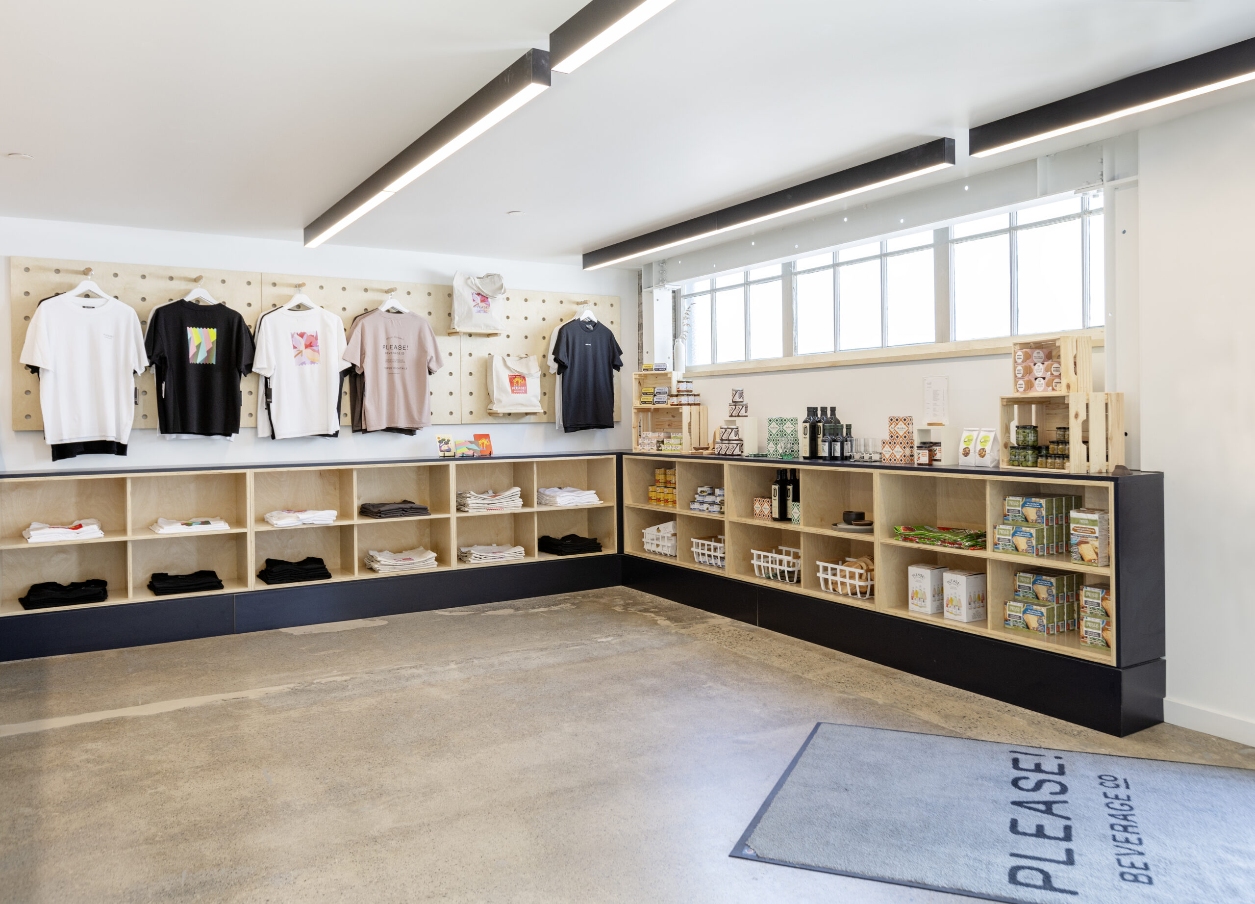 Bright and modern retail space within a beverage company featuring an array of merchandise including t-shirts displayed on a pegboard wall, and shelves neatly stocked with gourmet food items, beverages, and assorted goods, under warm ambient lighting with large windows providing natural light.