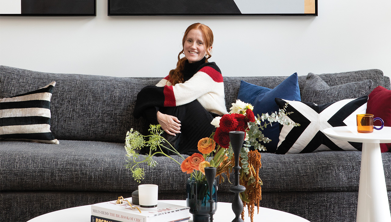 The designer sitting on a grey couch with vases of flowers and magazines on a coffee table in front.