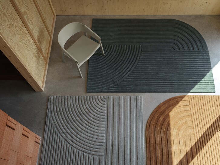 Above shot of three scuptural rugs (dark blue, grey and light brown) on the floor of a room with a white chair. 