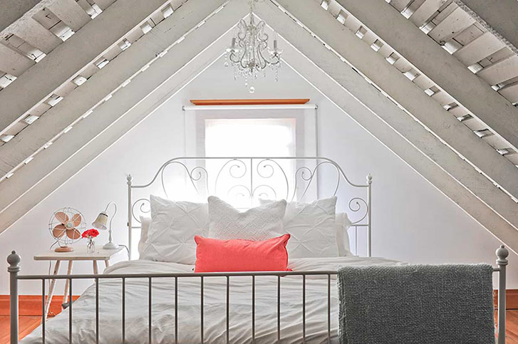 Crisp white bedding and a coat of eggshell paint on the wood beams keep this intimate space feeling bright and fresh.The peaked ceiling of the cozy bedroom loft is accentuated by a sparkling glass chandelier from The Cross Decor and Design. 